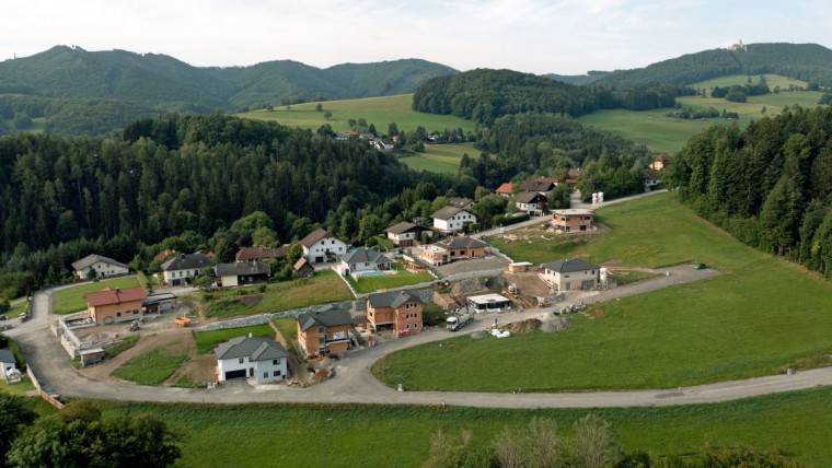 Die Siedlung Sonnenhang ist am Bild abgebildet. Zu sehen sind im Vordergrund fertige Häuser, Bauarbeiten an zu finalisierende Häuser. Das Bild aus Vogelperspektive bietet einen Ausblick an den umliegenden Wäldern und Hügeln.